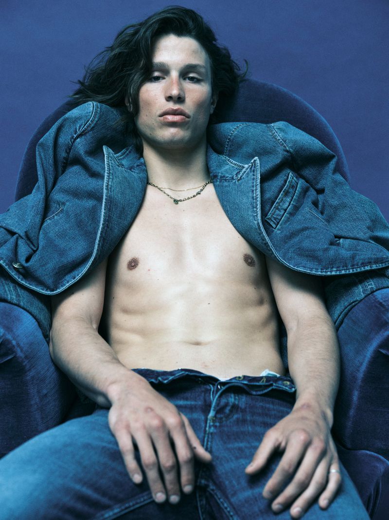 Shirtless man wearing an open denim jacket and blue jeans, seated in a blue velvet armchair against a solid blue background.