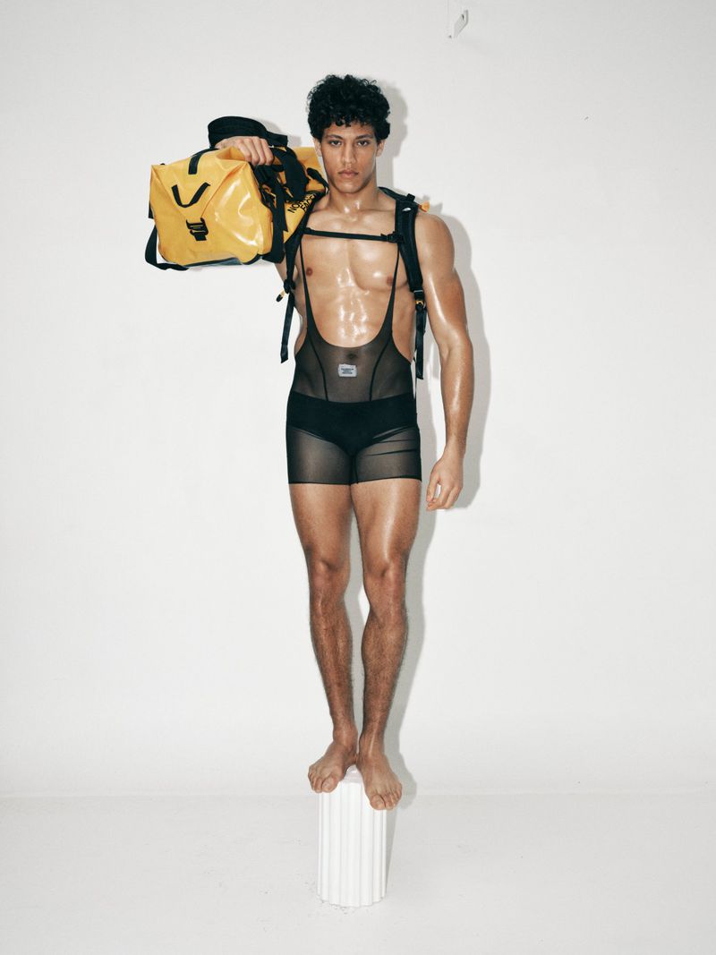 Model Lucas Barski standing on a white pedestal holding a yellow duffel bag in sheer black athletic wear against a white backdrop.