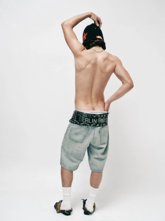 Shirtless man seen from behind wearing low-rise denim shorts, white socks, and a black beanie against a white studio background.