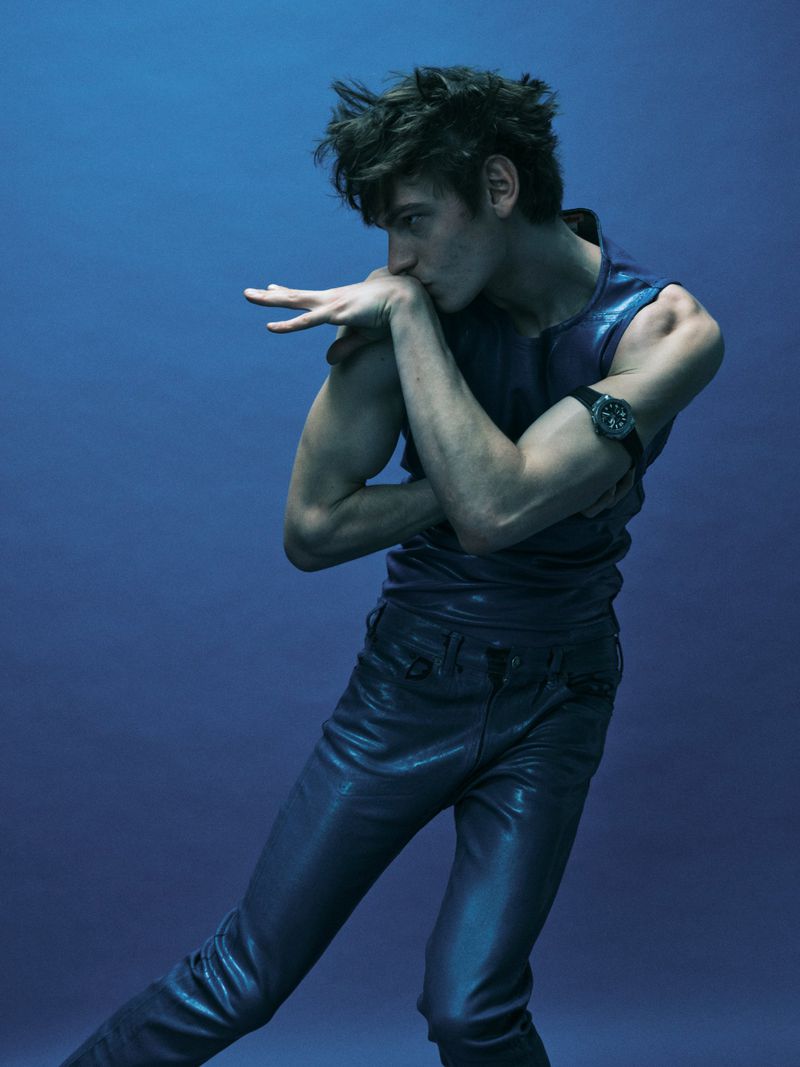 Model wearing dark blue leather trousers and sleeveless top posing against a blue studio background
