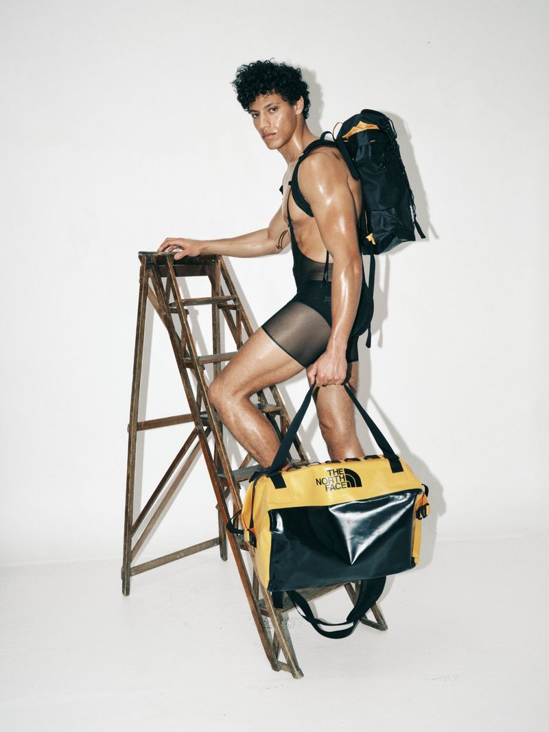Model Lucas Barski in sheer black shorts standing on a wooden ladder holding a yellow and black duffel bag against a white backdrop.