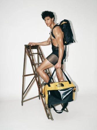 Model Lucas Barski in sheer black shorts standing on a wooden ladder holding a yellow and black duffel bag against a white backdrop.