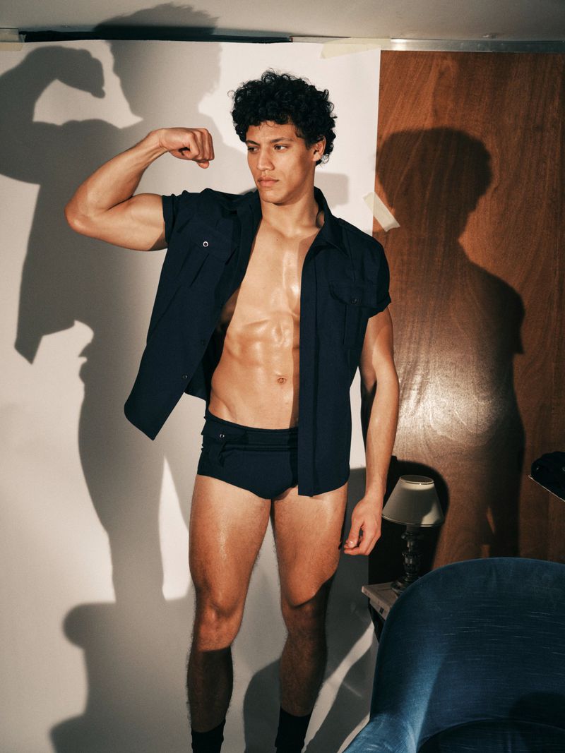 Model Lucas Barski posing shirtless in an open dark navy shirt and briefs while flexing against a studio set.