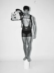 Model Lucas Barski balancing on a pillar, wearing a harness and holding a duffel bag against a white backdrop.