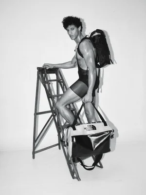 Black and white editorial photo of a man in a sheer bodysuit standing on a ladder holding a duffel bag against a white background.