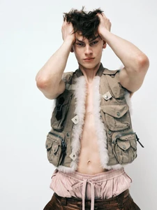 Model Lucas Barski wearing an open shearling-trimmed utility vest with hands in his hair against a white background.
