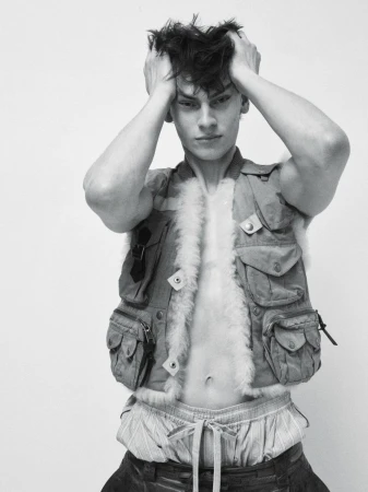 Model Lucas Barski posing with hands in hair wearing an open fleece-trimmed utility vest against a white studio background.