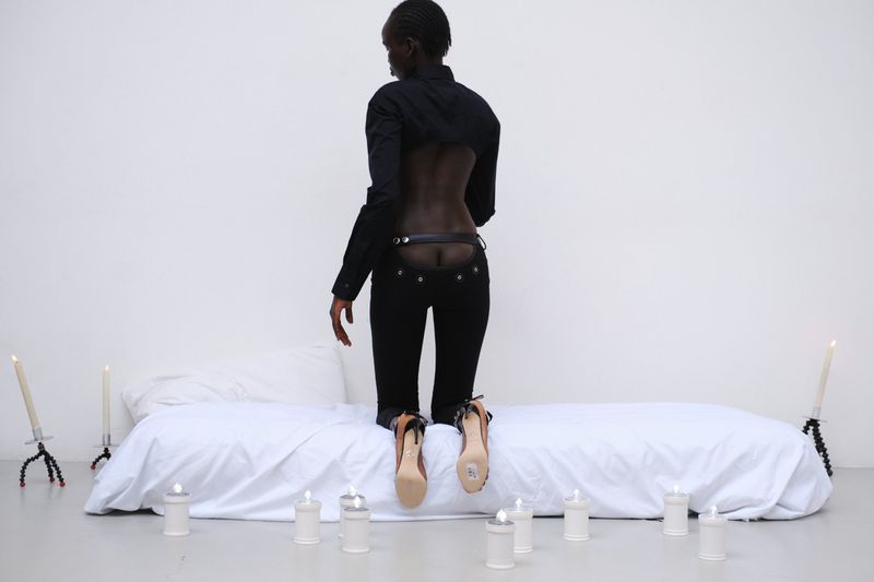 Model kneeling on a white mattress with back turned, wearing black backless attire and heels surrounded by lit candles in a white studio.