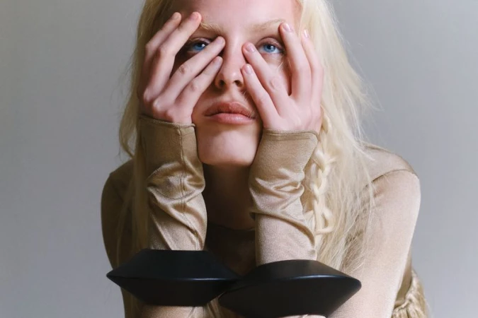 Close-up of a blonde model with hands framing her face, wearing a shimmering beige long-sleeve top and sculptural black cuffs.