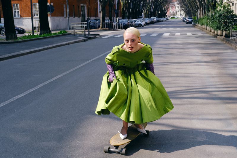 A woman wearing a voluminous lime green dress and purple gloves rides a skateboard down an empty street in white pointed heels.