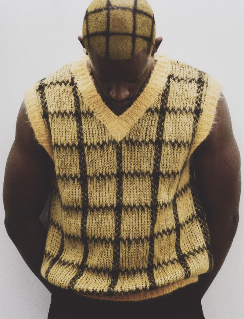Man with grid-patterned dyed hair looking down while wearing a matching yellow and black plaid knit sweater vest against a white backdrop.