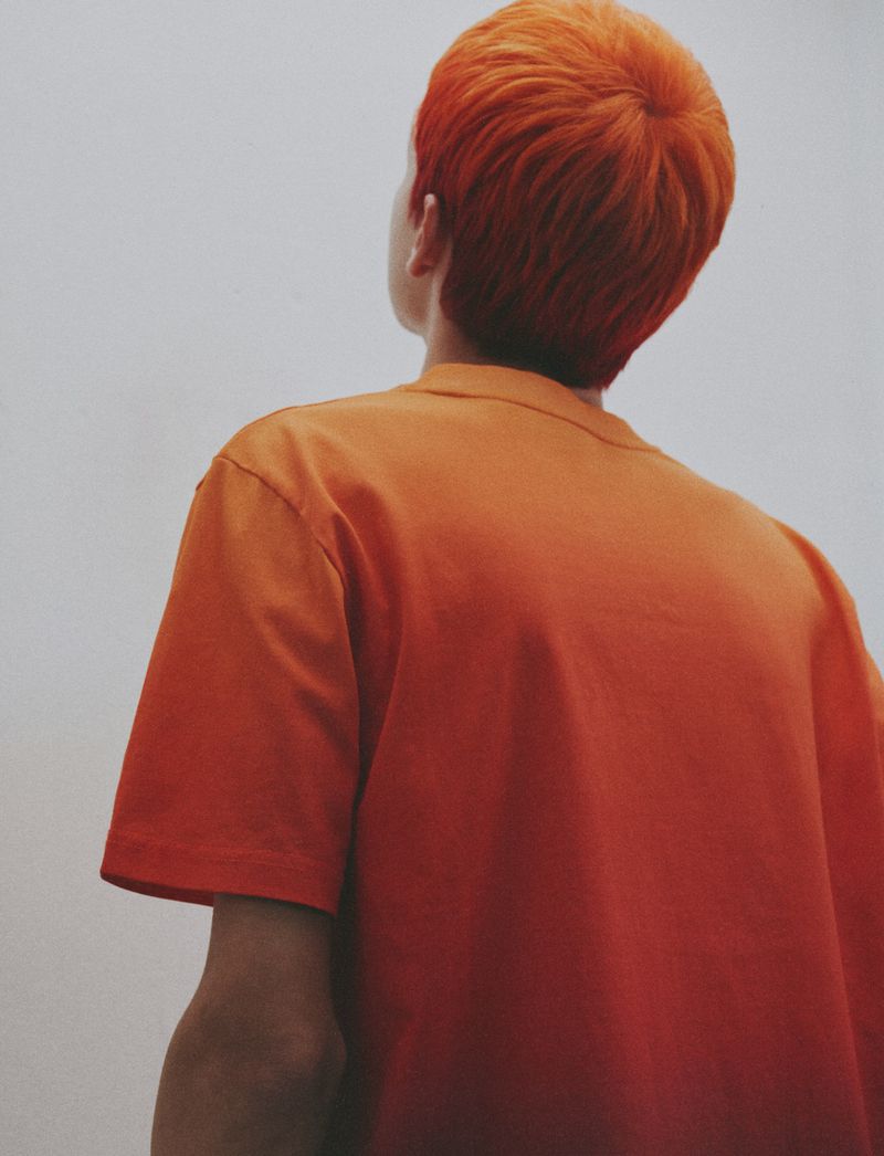 Back view of a person with vibrant orange hair wearing an orange t-shirt looking upward against a pale studio backdrop.