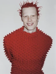 Smiling model with spiky red hair wearing a sculptural red popcorn textured top against a white studio background.