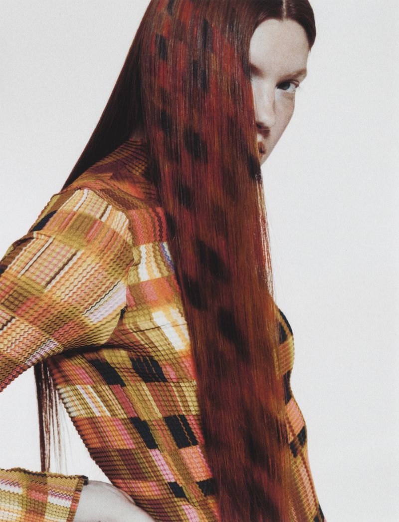Close-up portrait of a woman with patterned reddish-brown hair wearing a textured plaid top against a white backdrop.