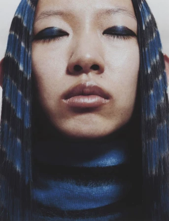Close-up portrait of an East Asian model with closed eyes, bold blue eye makeup, and blue and black patterned striped hair.