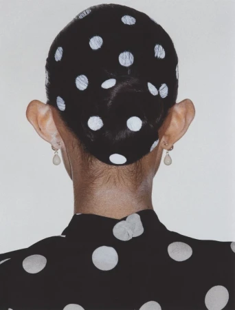 Back view of a person with hair in a bun painted with white polka dots, wearing matching black polka dot clothing against a white backdrop.