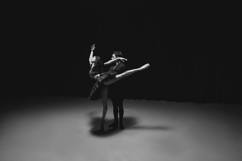 Monochrome photograph of a female ballet dancer in a black tutu supported by a male dancer in a dramatic pool of light.