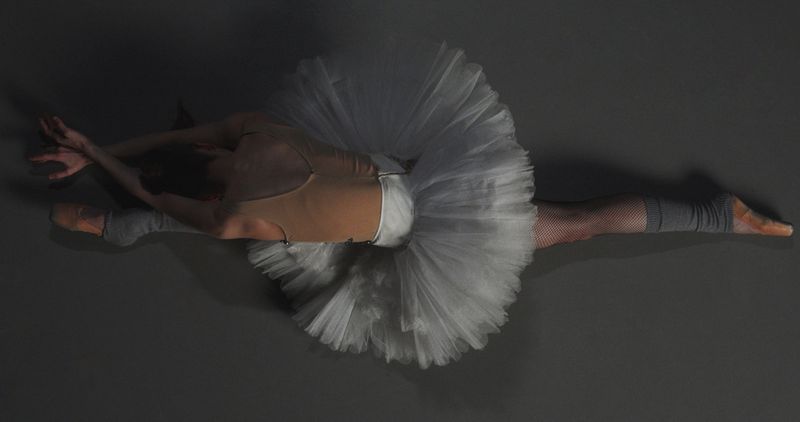 Overhead view of a ballerina stretching forward in full leg splits wearing a white tulle tutu and nude leotard on a dark grey floor.