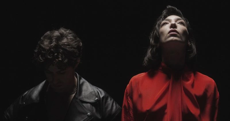 A woman in a vibrant red blouse looking upward alongside a man in a black leather jacket against a dark black background.