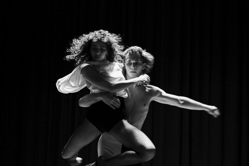 Black and white photo of a shirtless male dancer lifting a female dancer in fishnets and a flowing top against a dark backdrop.