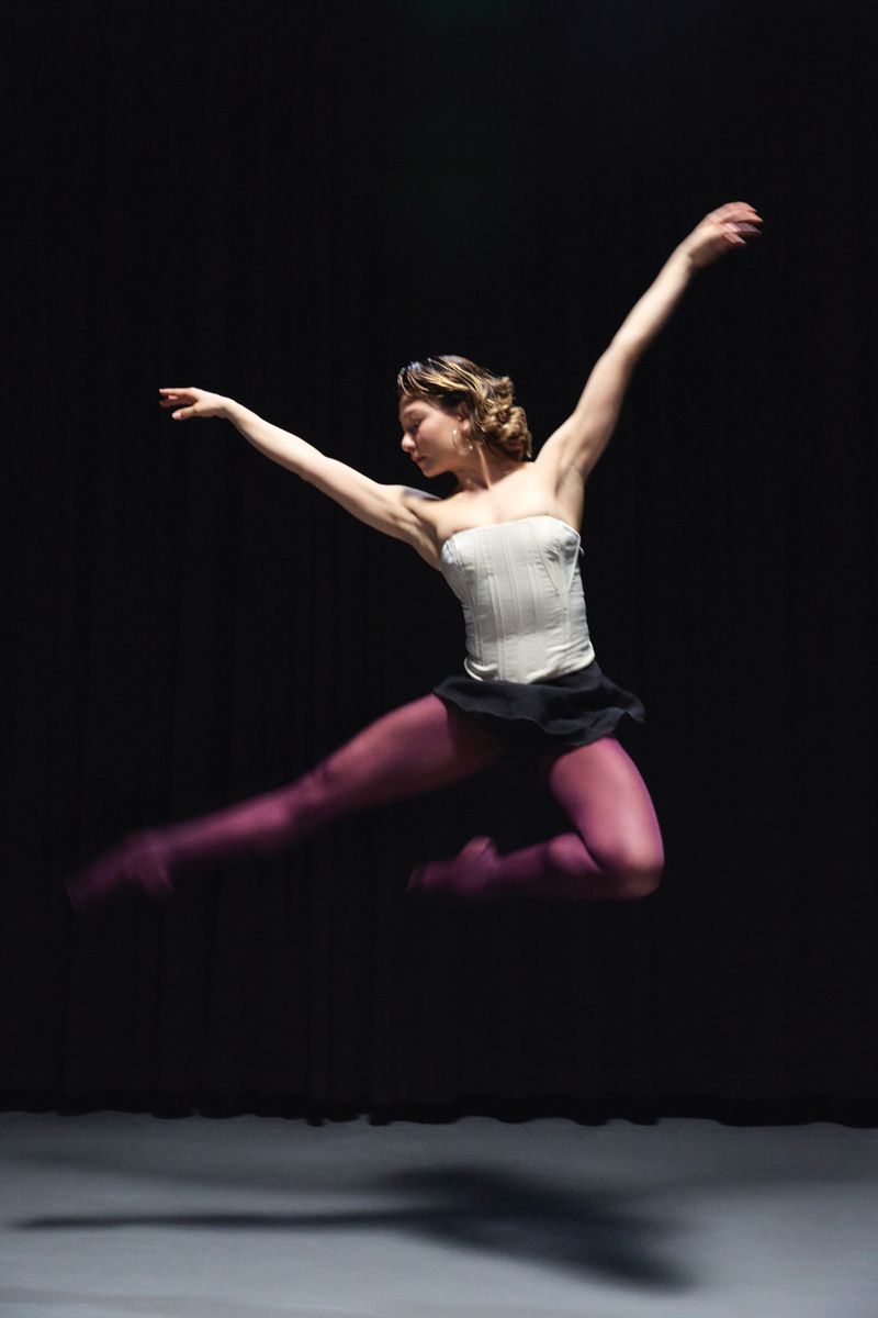 Ballerina leaping mid-air wearing a white corset, black skirt, and purple tights against a dark studio background.