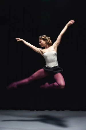 Ballerina leaping mid-air wearing a white corset, black skirt, and purple tights against a dark studio background.
