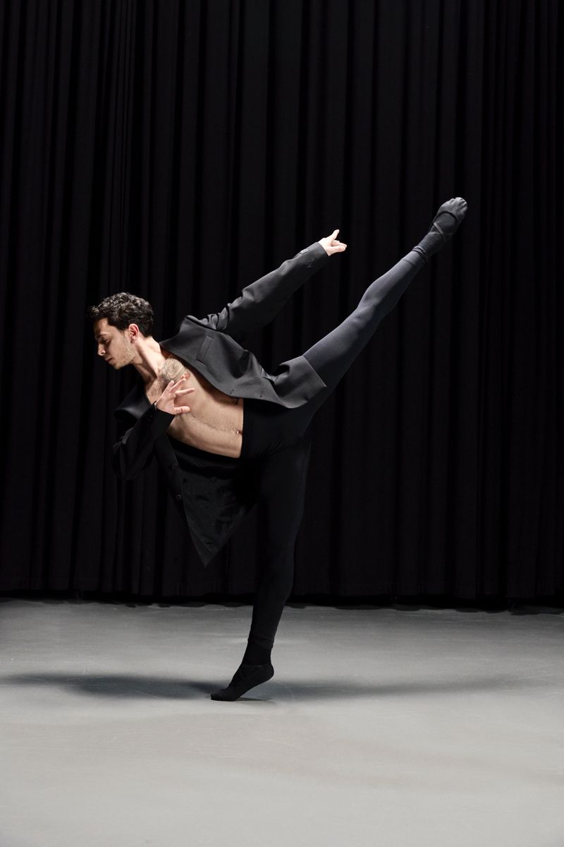 Male ballet dancer in dynamic extension wearing an open black blazer and black tights against dark curtains.