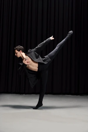 Male ballet dancer in dynamic extension wearing an open black blazer and black tights against dark curtains.
