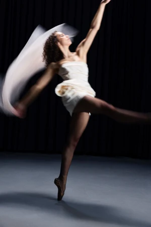A ballet dancer in a strapless white dress dances en pointe against a dark studio background with motion blur on fabric.