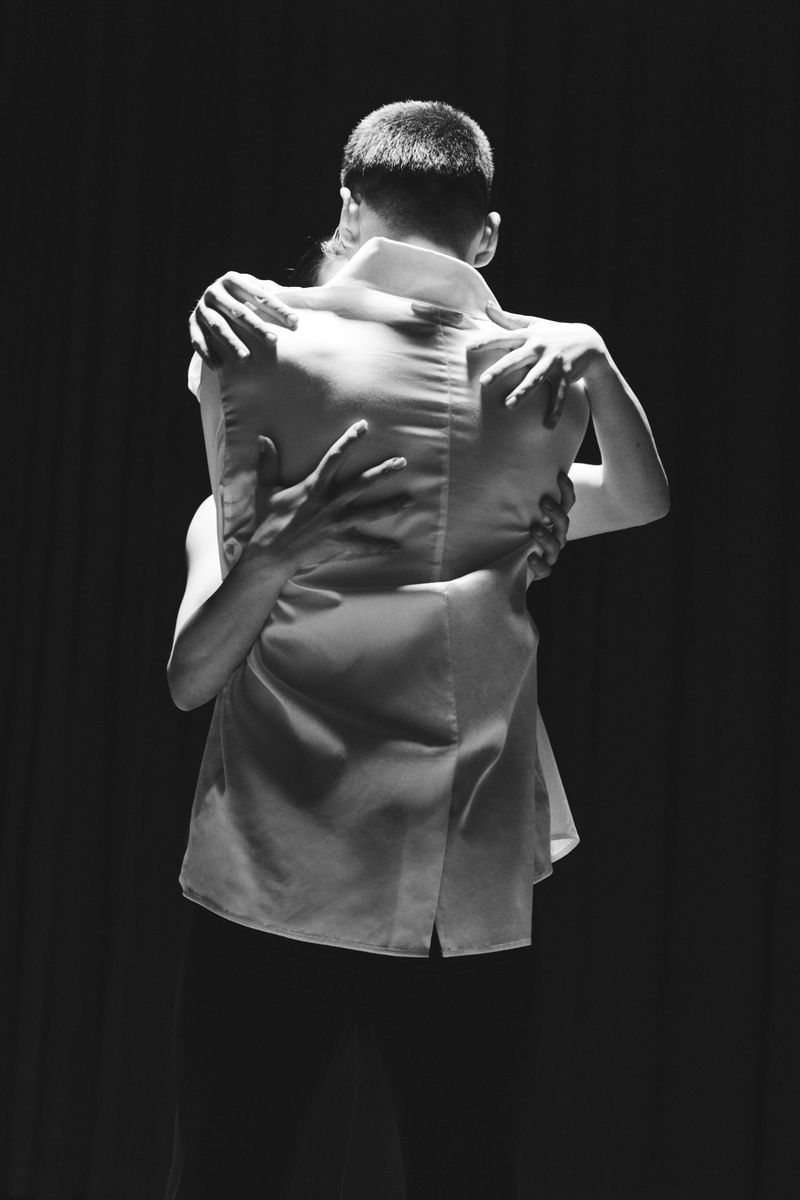 Black and white photograph of two people closely embracing, viewed from behind as arms wrap around a white collared vest.