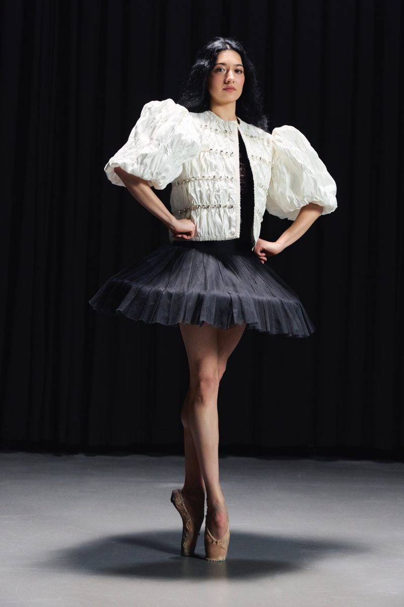 Ballerina in a white puff-sleeve jacket, black tutu, and pointe shoes posing en pointe against a dark backdrop.