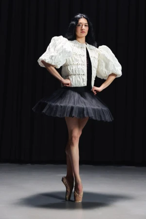 Ballerina in a white puff-sleeve jacket, black tutu, and pointe shoes posing en pointe against a dark backdrop.