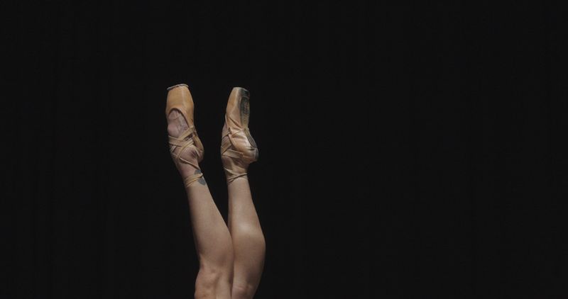 Ballet dancer legs wearing satin pointe shoes extended upward against a solid black background.