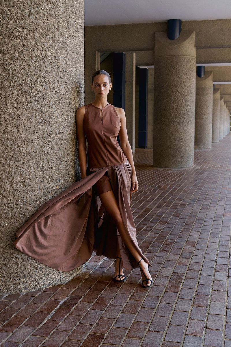 Model leaning against a massive concrete pillar in a brown sleeveless dress with a flowing pleated overlay skirt.
