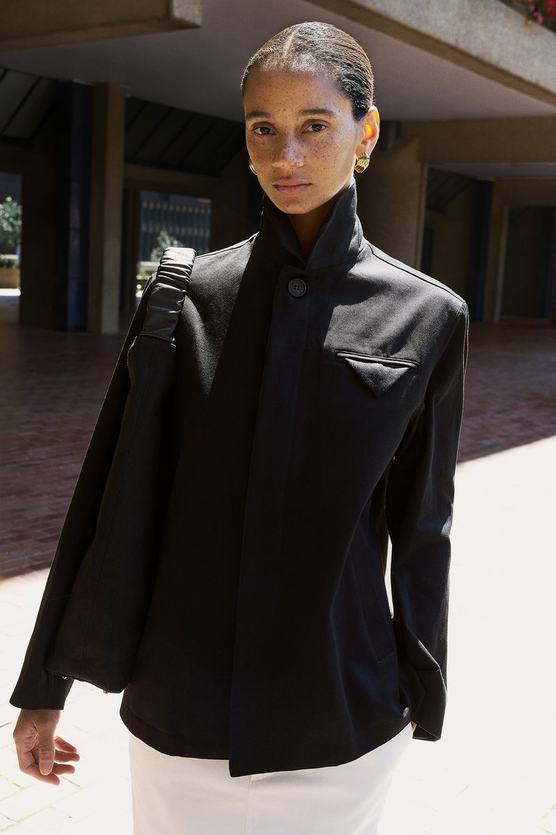 Model wearing a structured high-collar black jacket and carrying a black shoulder bag outdoors in bright sunlight.