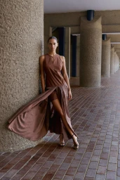 Model leaning against a massive concrete pillar in a brown sleeveless dress with a flowing pleated overlay skirt.