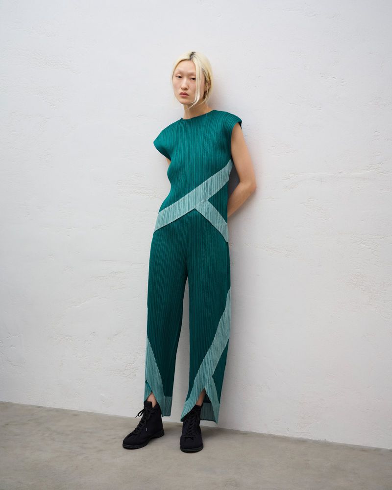 Model in a green sleeveless pleated jumpsuit with geometric mint stripes and black lace-up boots leaning against a pale textured wall.