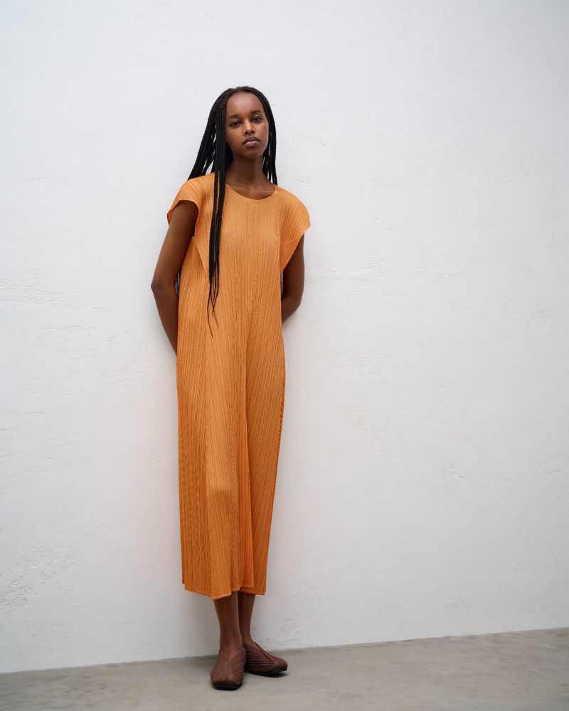 Black woman standing against a white wall wearing an orange pleated sleeveless dress and brown shoes.