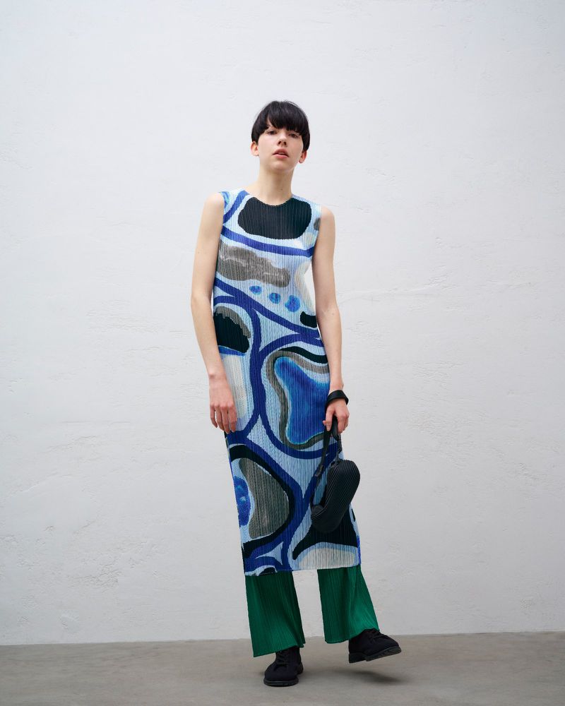 Model wearing a blue printed pleated dress over green trousers against a plain textured studio wall.