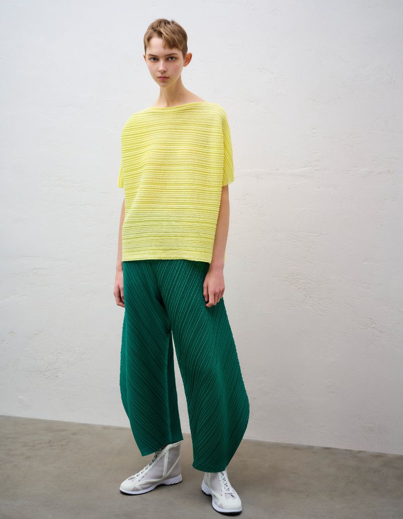 Model wearing a pleated yellow top and emerald green wide-leg trousers with white sneakers against a neutral studio wall.