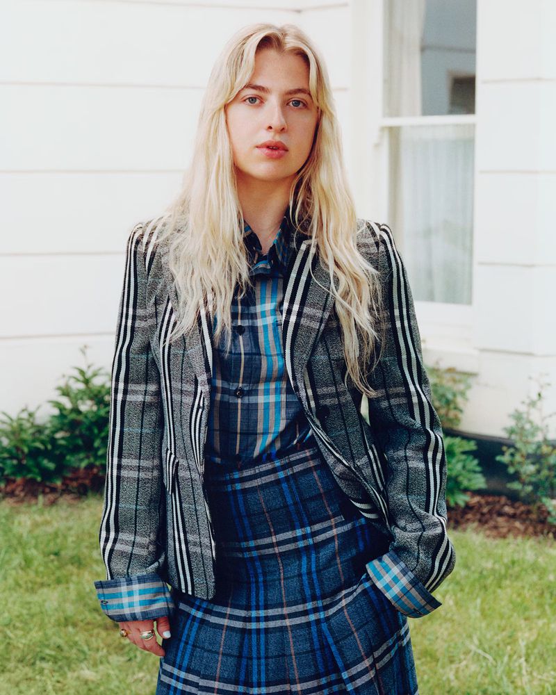 Woman in stylish grey plaid suit and skirt posing outdoors near a white building; blonde hair, medium shot, eye-level perspective, natural lighting.