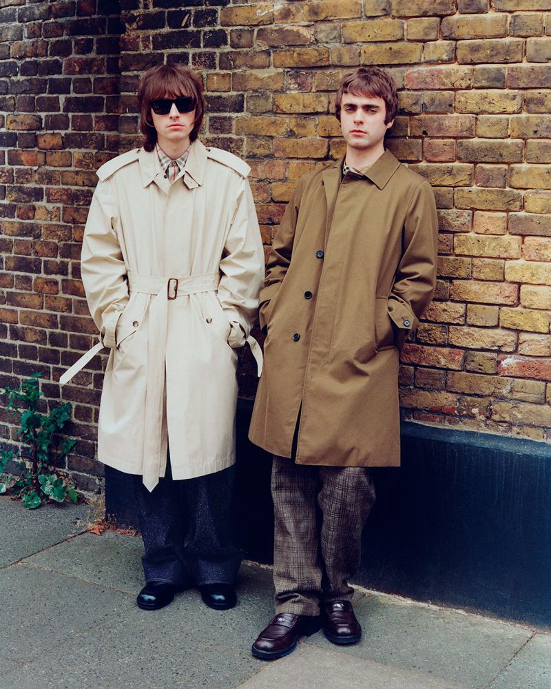 Two adult men pose against a brick wall in khaki trench coats and trousers, captured in a medium shot with natural lighting.