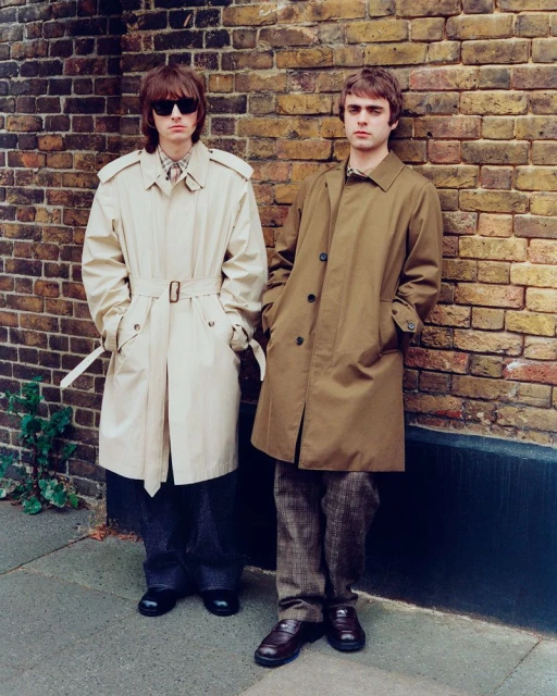 Two adult men pose against a brick wall in khaki trench coats and trousers, captured in a medium shot with natural lighting.
