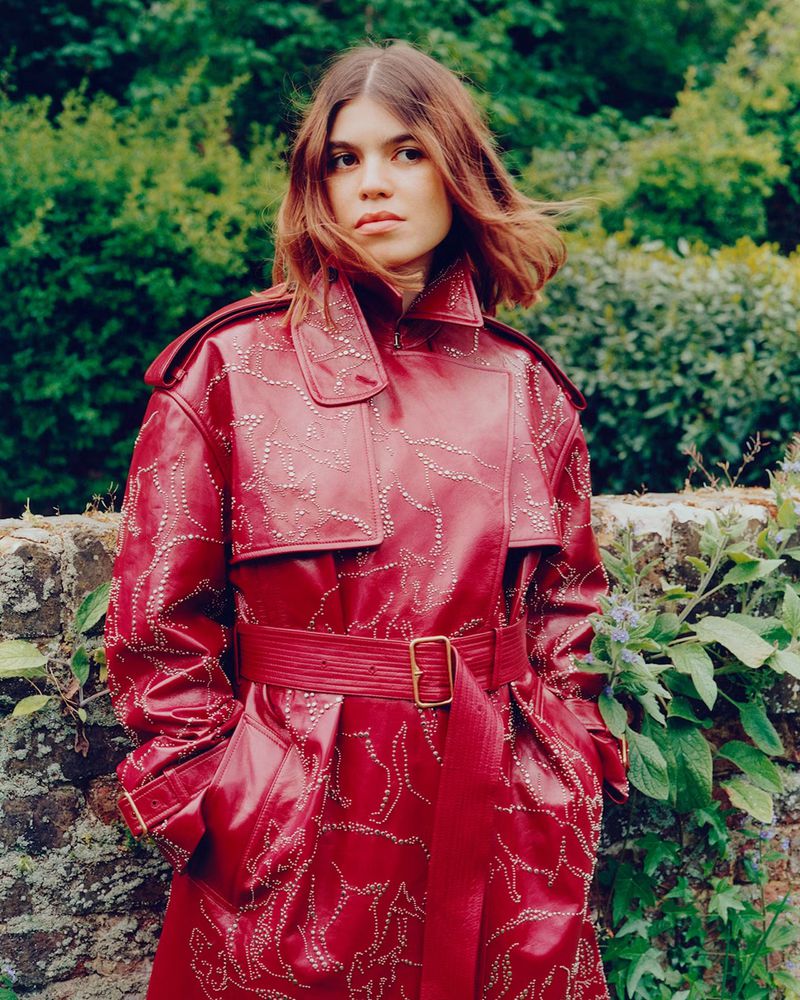 An adult woman with brown hair is shown wearing a red leather coat with embellishments standing in front of a stone wall and greenery.