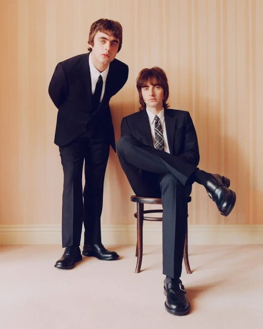 Two men in black suits pose indoors; one seated on a wooden chair, both looking at the camera against a peach striped wall.