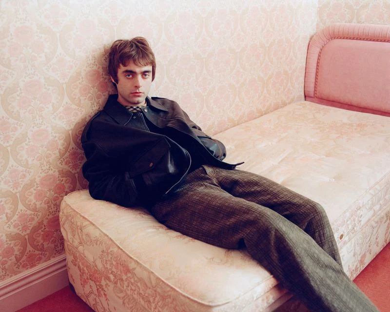 Young man in a black leather jacket and plaid pants poses indoors leaning against a floral patterned wall and bed frame.
