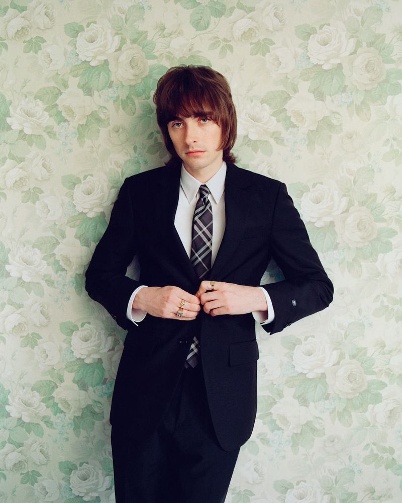 Portrait of a dark-haired adult man wearing a dark suit and tie against a floral wallpaper backdrop in an indoor setting.