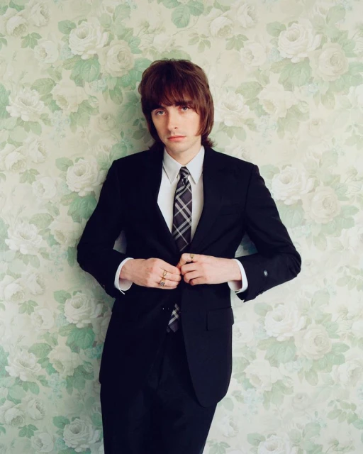 Portrait of a dark-haired adult man wearing a dark suit and tie against a floral wallpaper backdrop in an indoor setting.