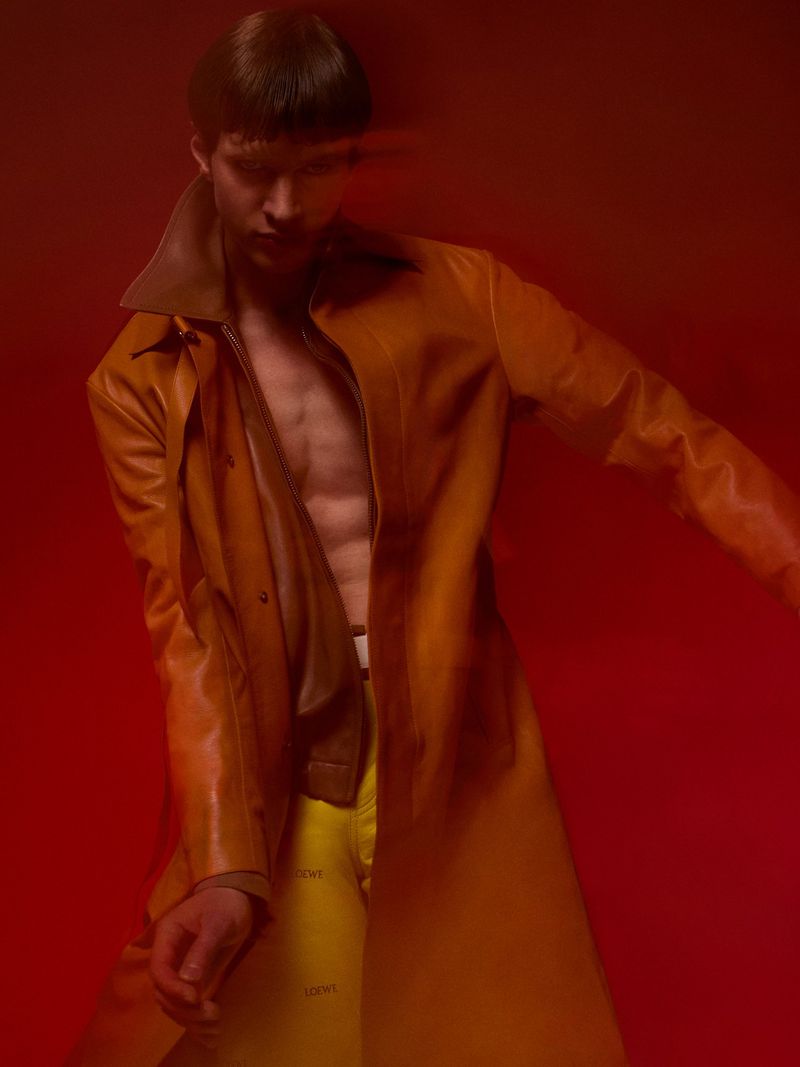 Male model Kris Wardak posing in an open tan leather coat and yellow trousers against a red seamless studio background.