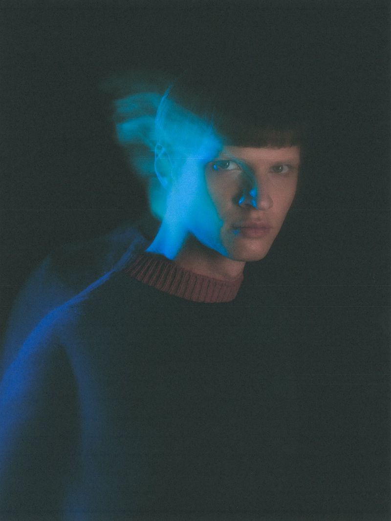 Model Kris Wardak in a dark ribbed collar sweater with blue motion blur streaks against a black background.